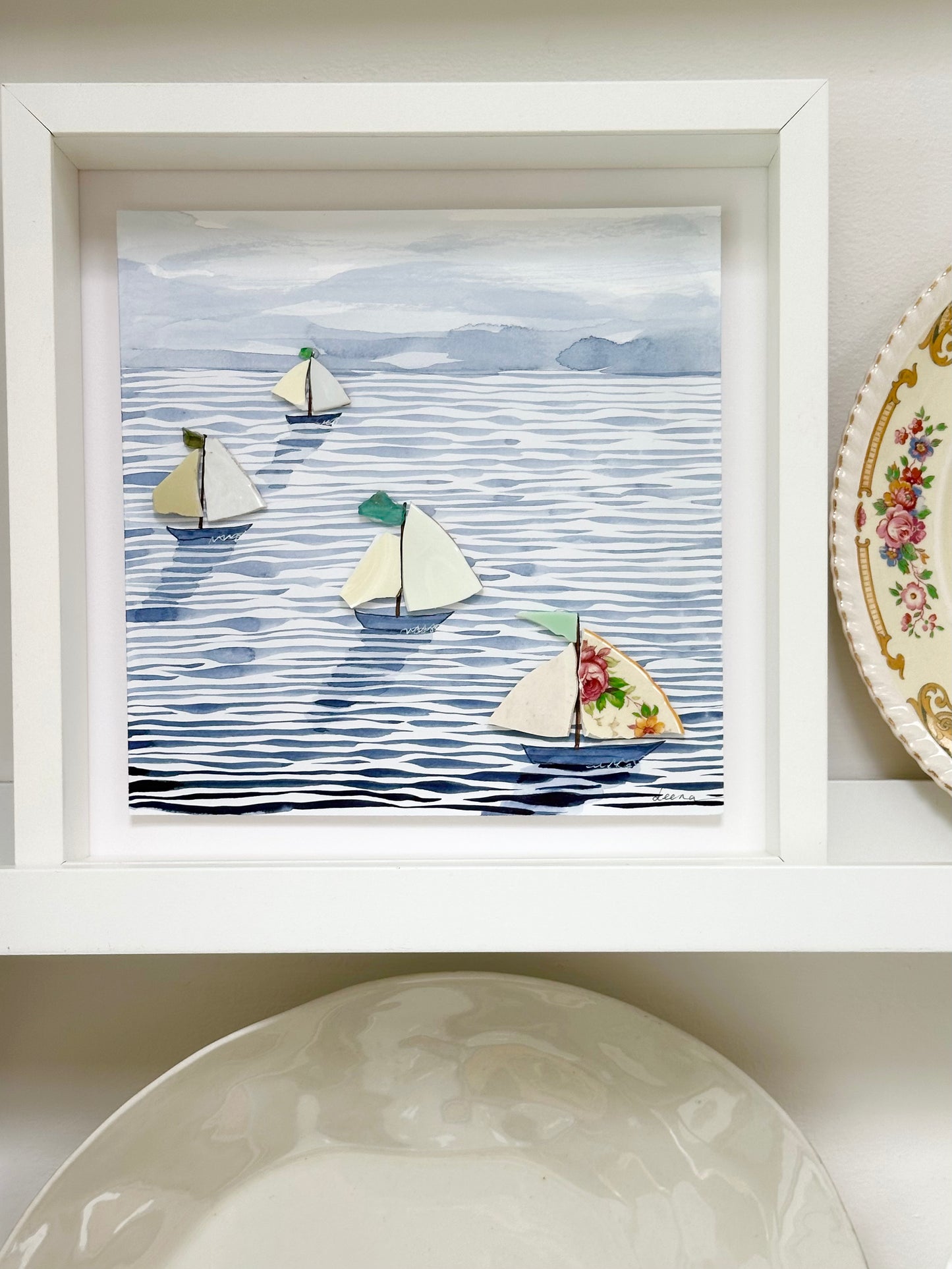 “Mixed media coastal artwork with watercolour waves and sailboats made from reclaimed china shards in a white frame.”b